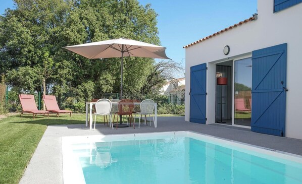 Una piscina con una mesa y sillas, una puerta azul y un edificio blanco con contraventanas azules.