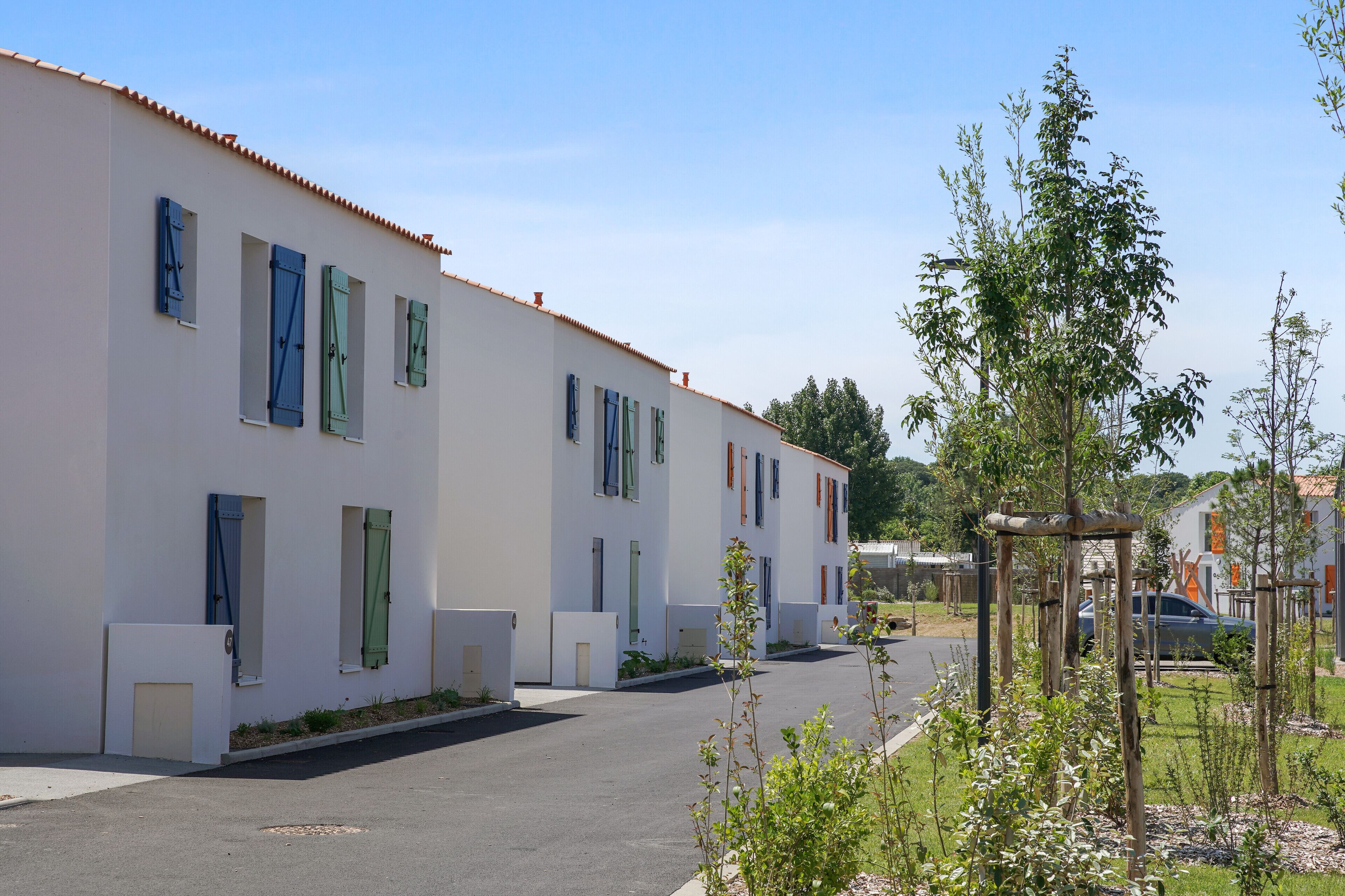 A row of white buildings with multiple windows and doors, a clear sky, and a parking area with a few cars.