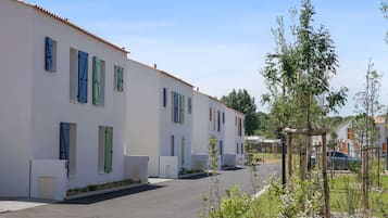 A row of white buildings with multiple windows and doors, a clear sky, and a parking area with a few cars.