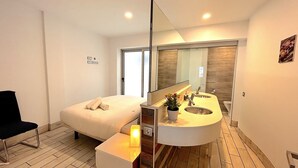 Ein modernes Hotelzimmer mit Bett, Nachttisch mit Lampe, großem Spiegel, Bad mit Waschbecken und Toilette sowie einer Topfpflanze.