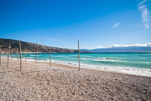 A pebble beach with wooden poles, a clear blue sea, and distant mountains.