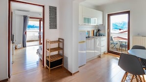 Una cucina compatta con mobili bianchi e un forno a microonde integrato. Contro il muro si trova un'unità a scaffali in legno, e si intravede un'area pranzo con tavolo e sedie.