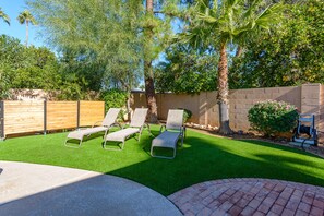 Un jardin avec une pelouse artificielle, deux chaises longues, un palmier et un mur en briques.