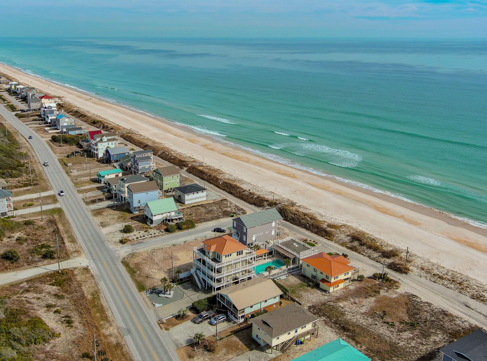 An aerial view of a coastal residential area with houses along the beach, a road, and a swimming pool in a building.