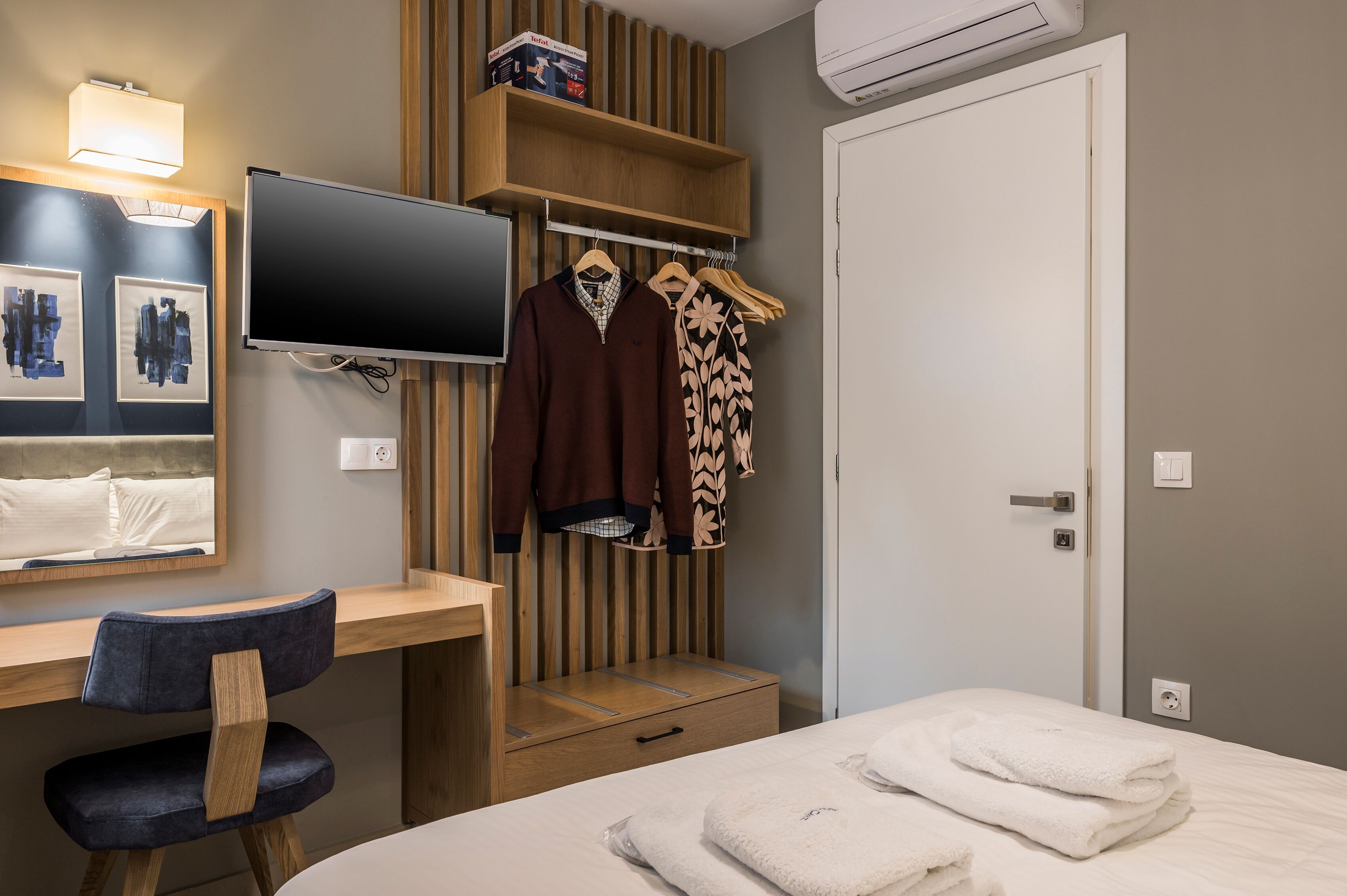 Una habitación de hotel con una cama, un escritorio con silla, un televisor, un armario con ropa y una estantería en la pared.