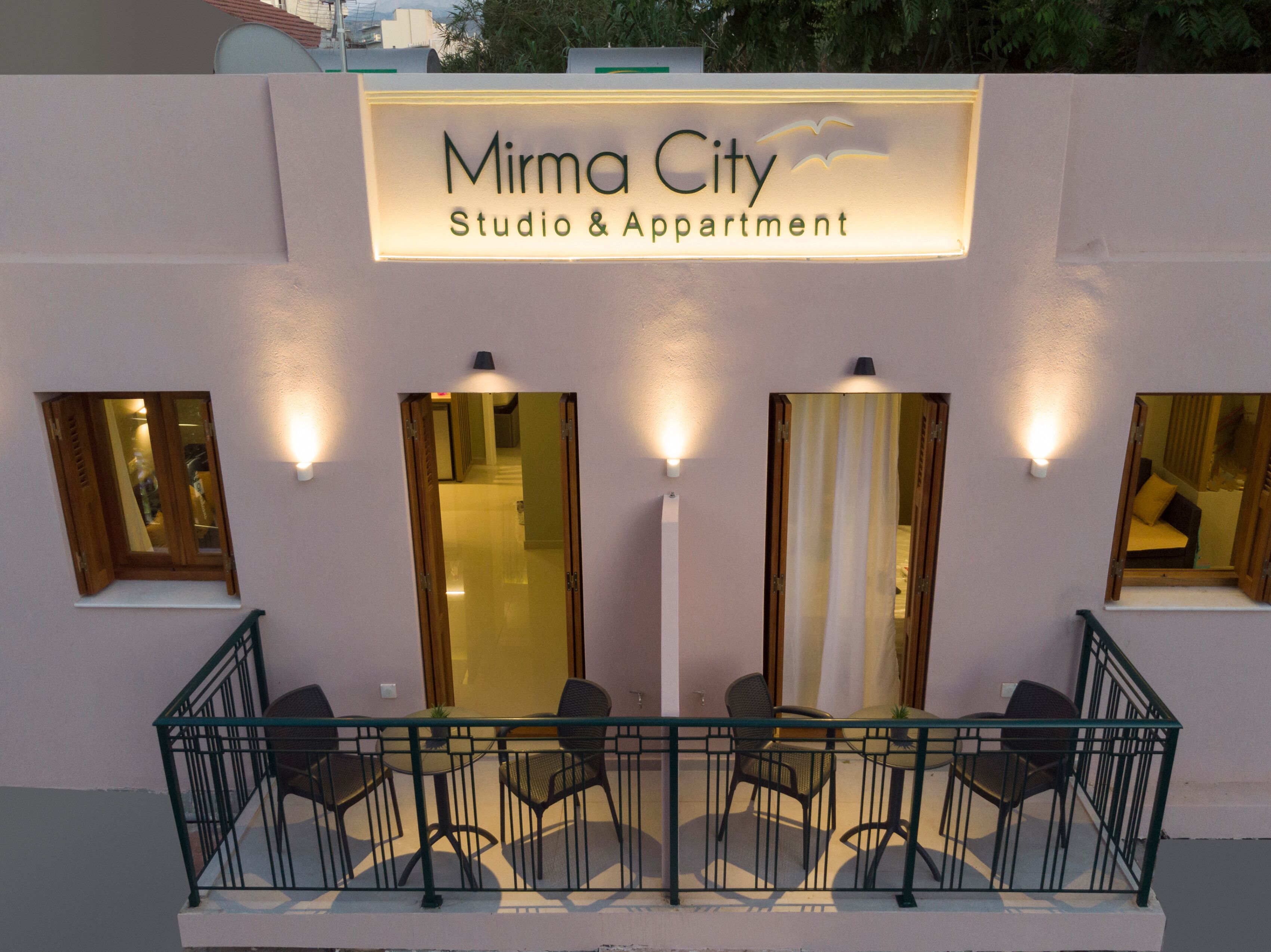 En bygning med et skilt, der viser "Mirma City Studio & Apartment". Fire vinduer med gardiner, en balkon med stole og gelænder.