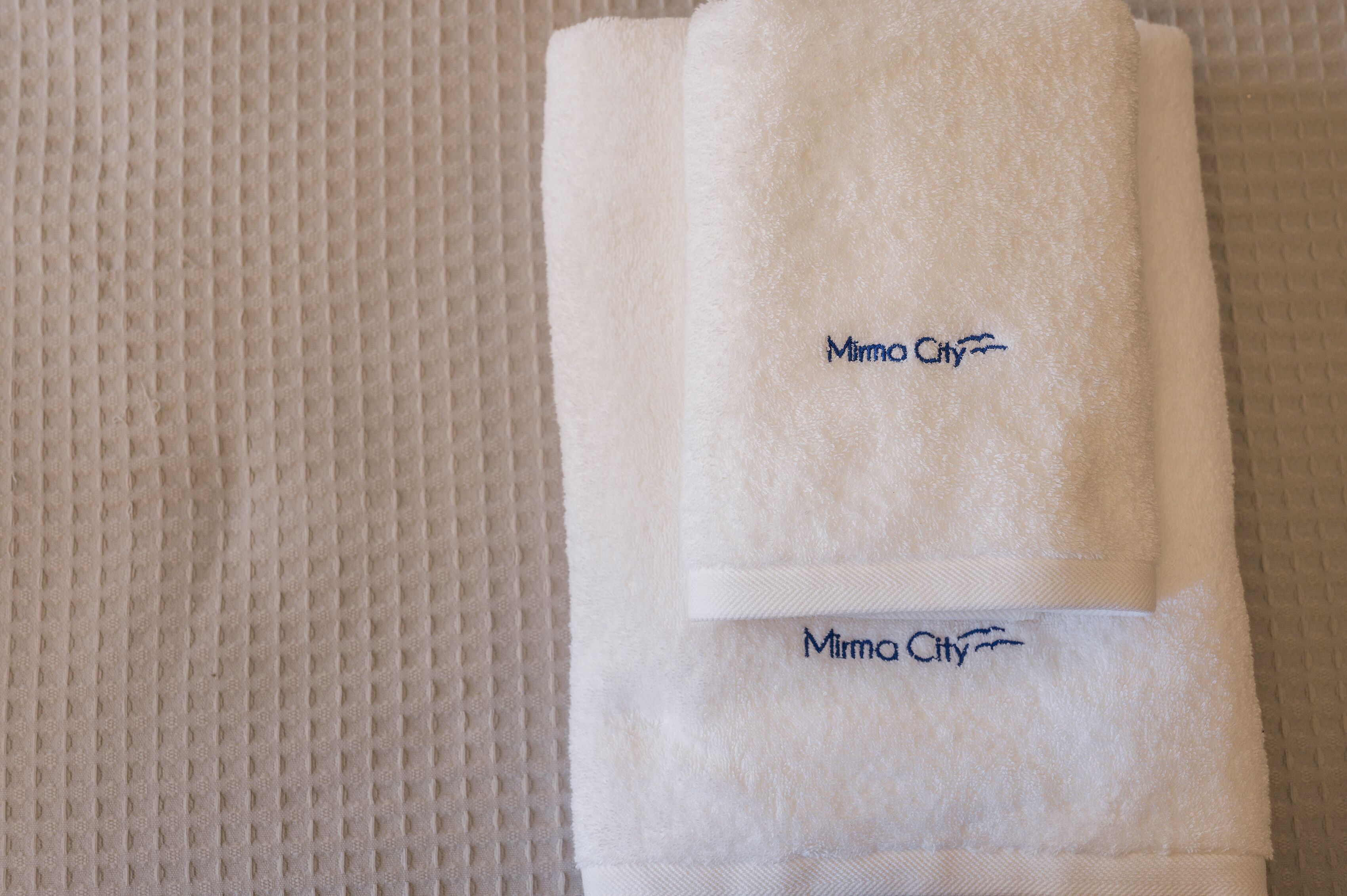Une serviette blanche pliée, ornée d’une broderie représentant le nom « Mima City », est posée sur une surface texturée.