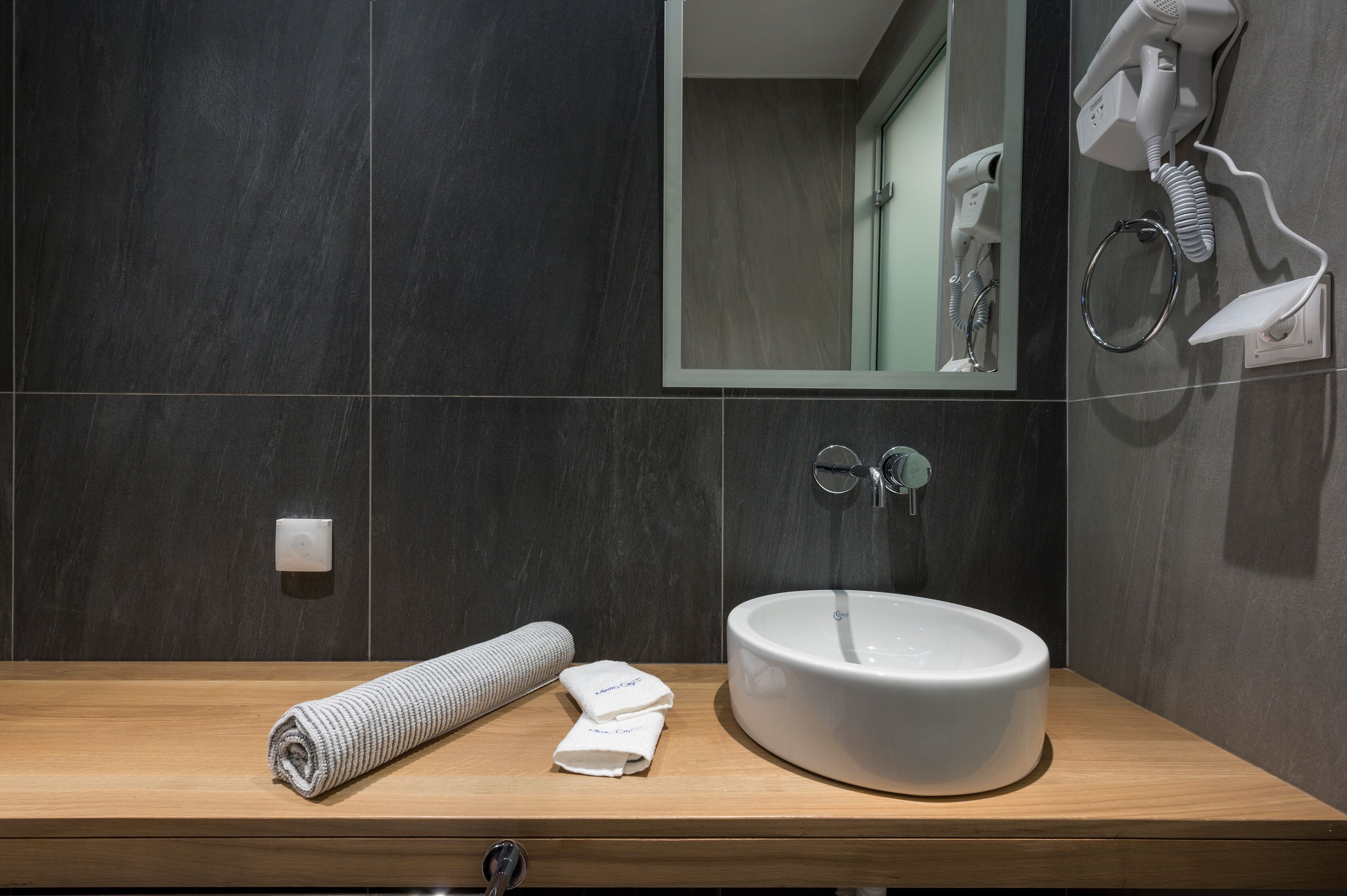 Une salle de bain moderne avec un meuble-lavabo en bois, un lavabo blanc, une serviette et un sèche-cheveux fixé au mur.