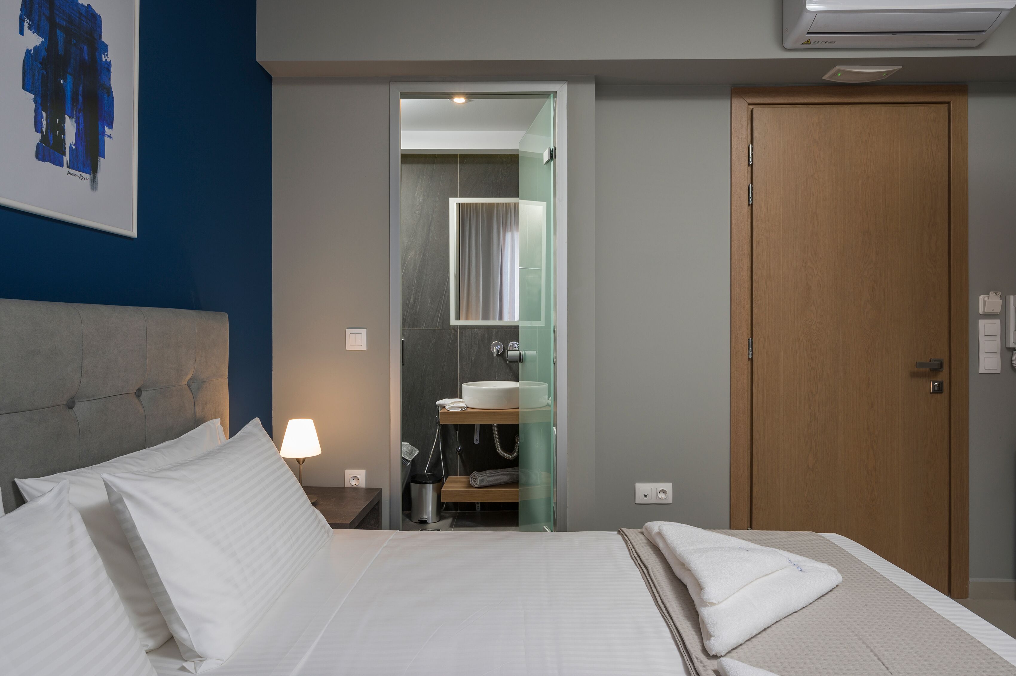 A hotel room with a bed, a nightstand with a lamp, a bathroom with a sink and mirror, and a wooden door.