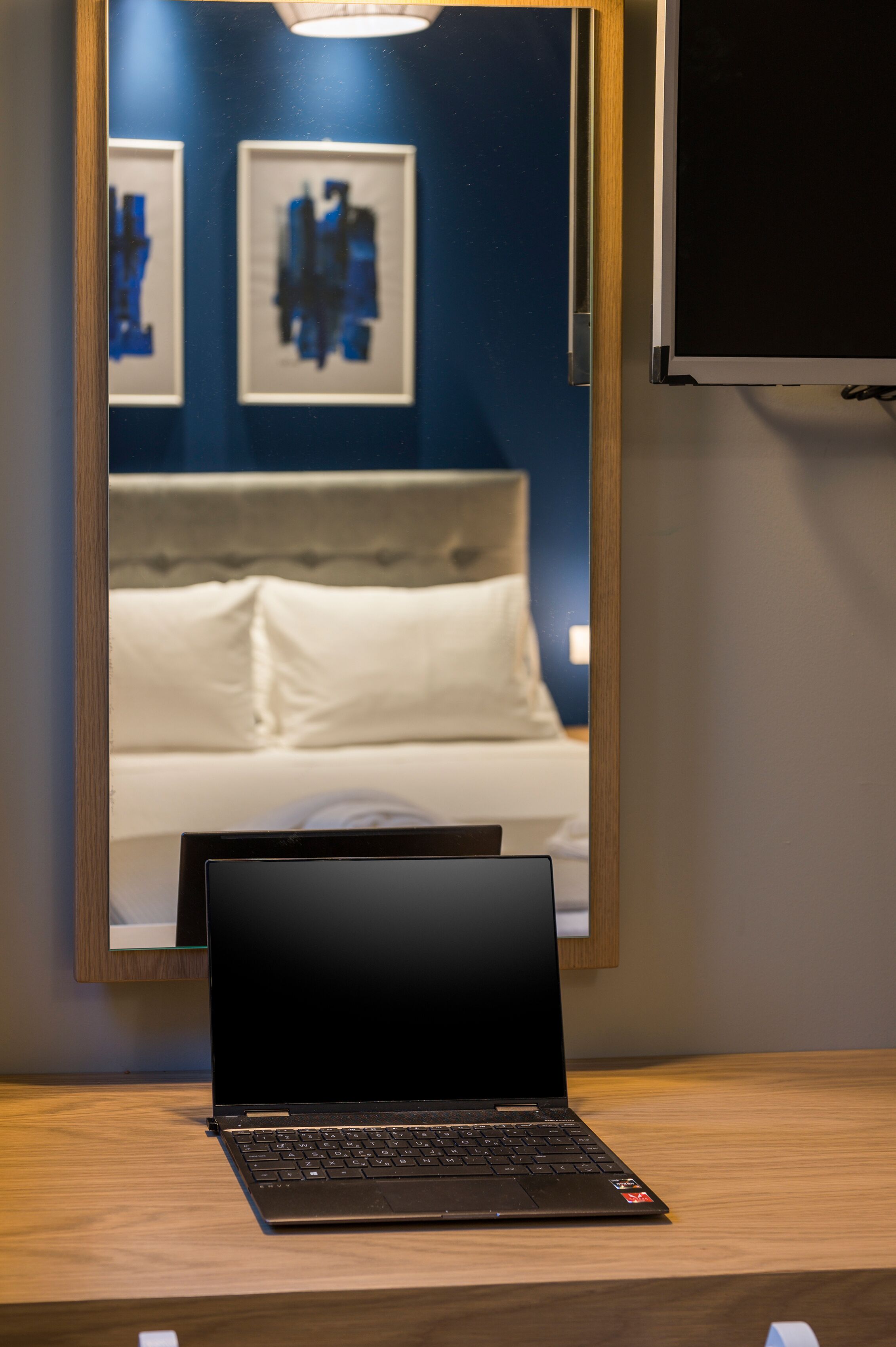 Ein Laptop auf einem Holztisch, ein Spiegel zeigt ein Bett und Kunstwerke.