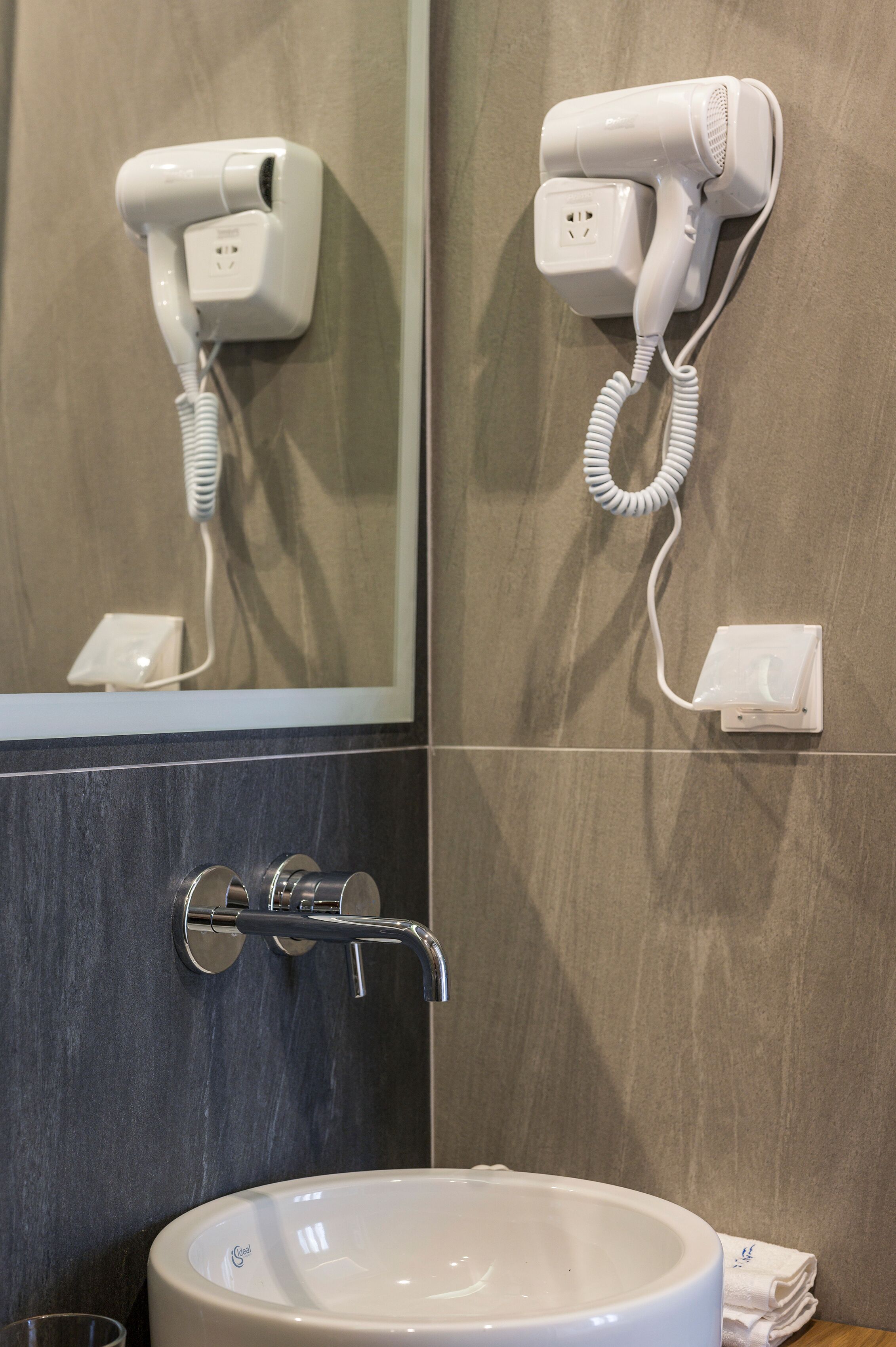 Un baño con un secador de pelo de pared, un lavamanos con grifo y un espejo que refleja el secador.