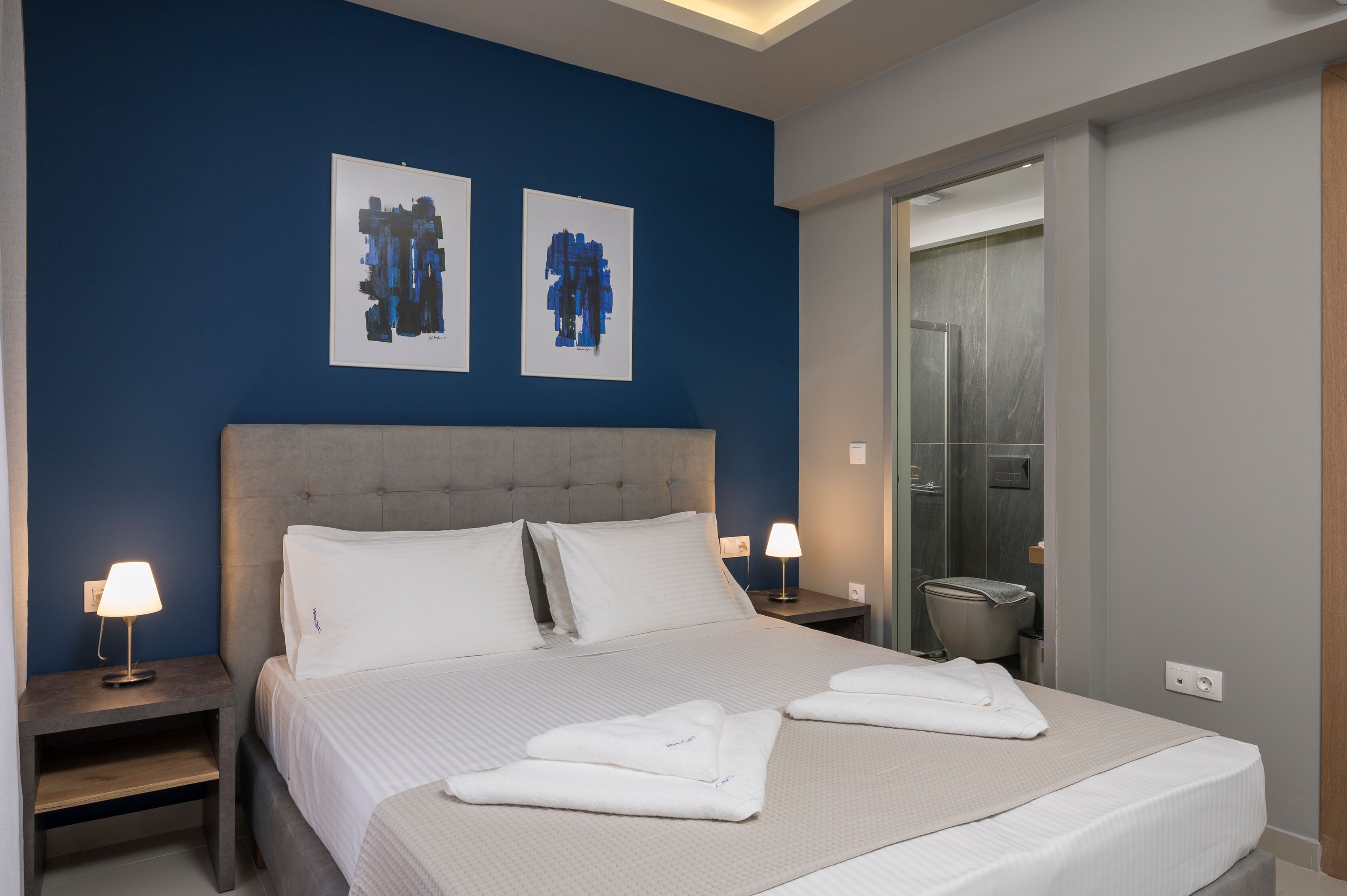 Une chambre d’hôtel avec un grand lit, deux lampes de chevet, une tête de lit grise et des œuvres d’art accrochées au mur bleu.