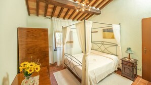 A bedroom with a four-poster bed, wooden ceiling beams, a bedside table with a lamp, and a vase of sunflowers.