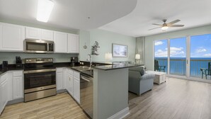 A modern kitchen with white cabinets, stainless steel appliances, and a wooden floor. There is a balcony with a view of the ocean. - Calypso 2003W | 20th-Floor Gulf Views, Beach Service Included, Steps from Pier Park (Panama City Beach)