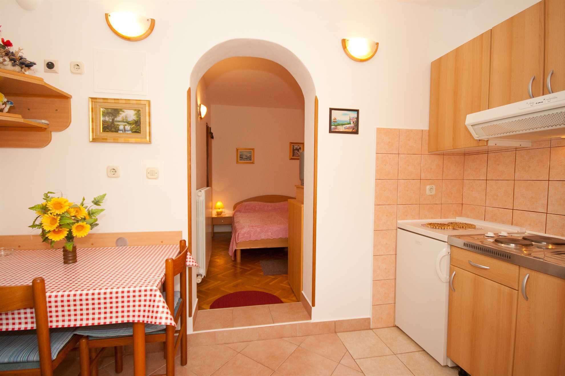 A compact kitchen with a stove, oven, and cabinets. There is a dining area with a table and chairs, and a bedroom visible through an archway.