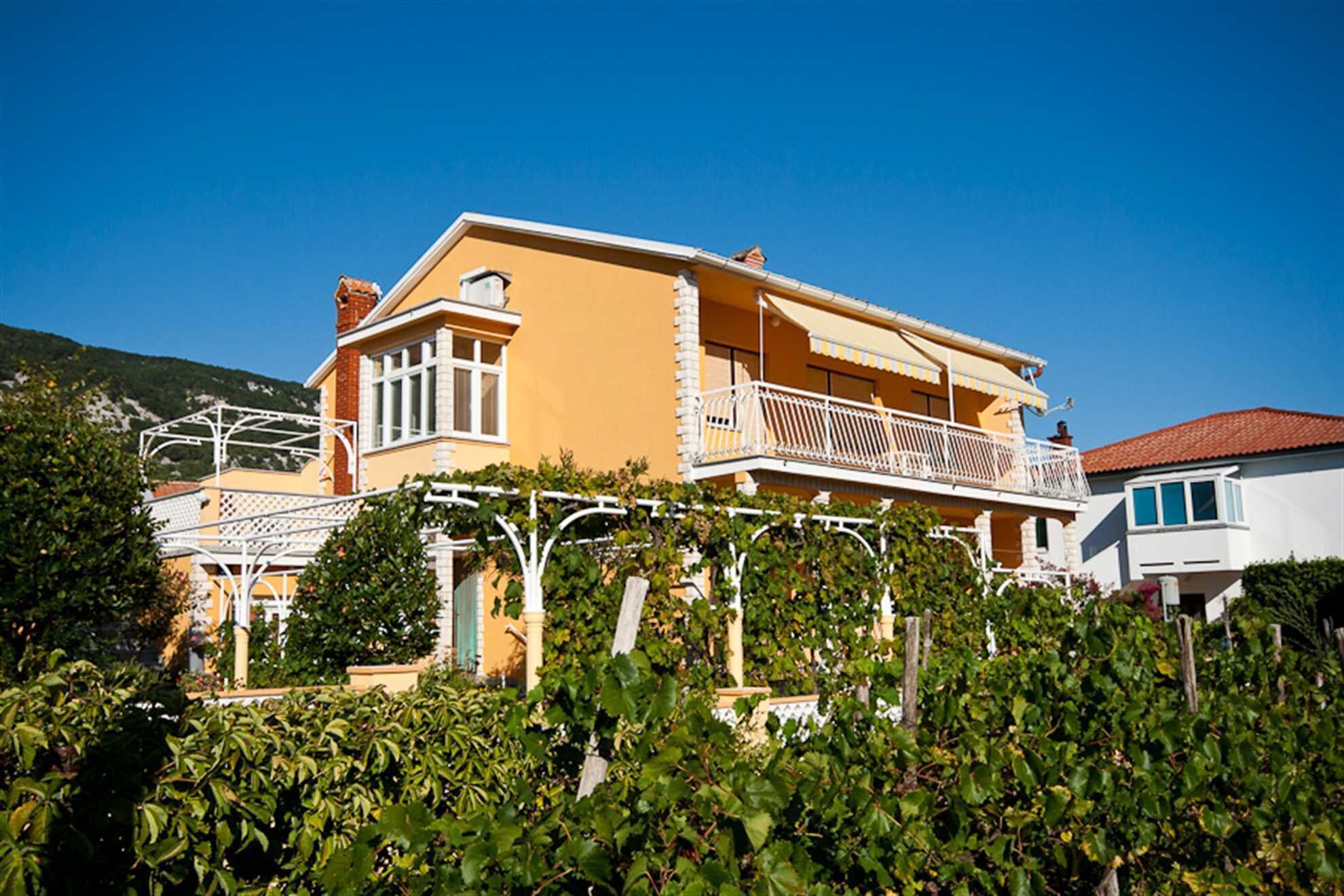 A two-story building with a balcony, surrounded by greenery and a clear blue sky.