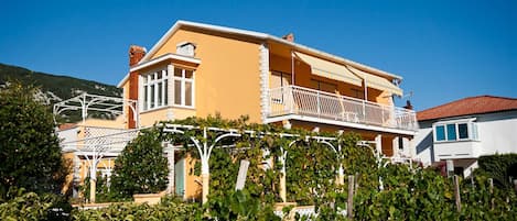 A two-story building with a balcony, surrounded by greenery and a clear blue sky.