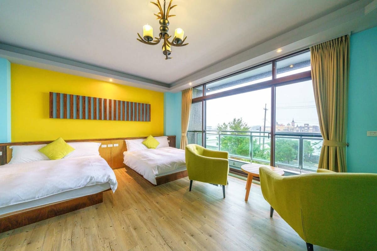 Quarto de hotel com duas camas, uma janela grande com vista, um candelabro e uma cadeira amarela.