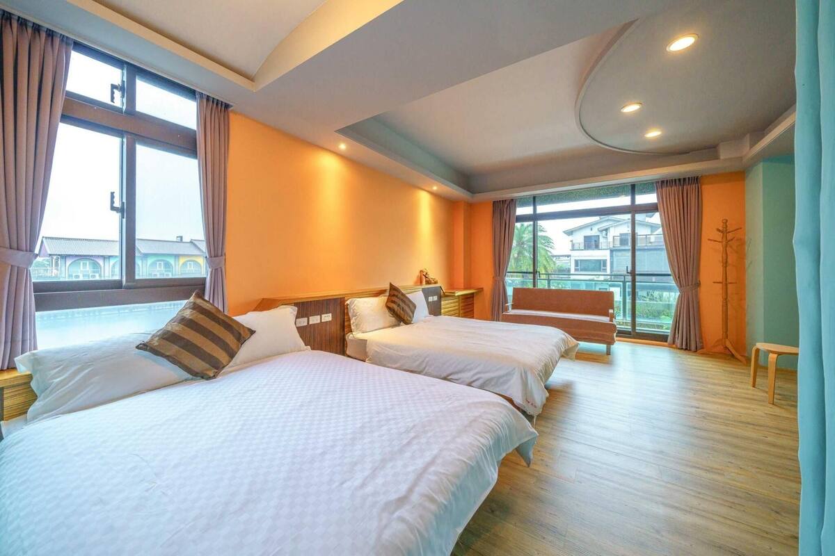 Quarto de hotel com duas camas, piso de madeira e janelas amplas com cortinas.