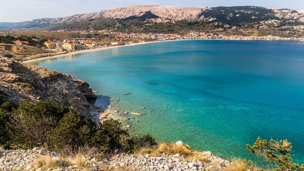 Un pueblo costero con aguas azul turquesa, acantilados rocosos y montañas al fondo.