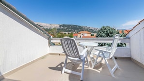 Un balcón con sillas de plástico blanco y una mesa redonda pequeña, con vistas a un paisaje montañoso con casas al fondo.