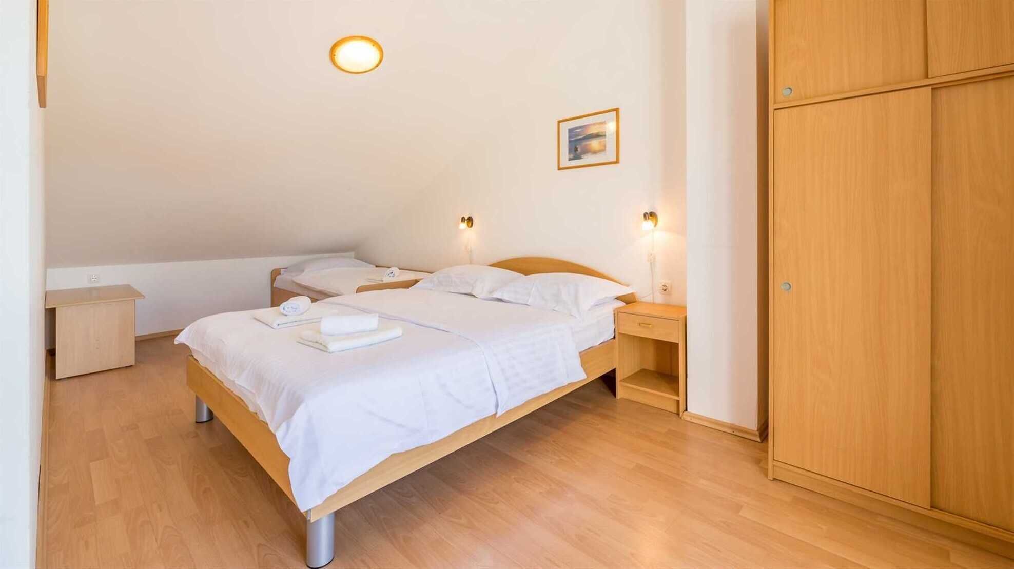 A single bed with white linens, two bedside tables, a nightstand, a wooden wardrobe, and two wall-mounted light fixtures.