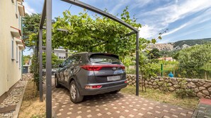 A Kia Sportage parked under a metal structure in a residential area with a paved driveway and stone wall. - Janezic apartment- Charming 2-Bedroom Apartment with Parking and grill (Baška)