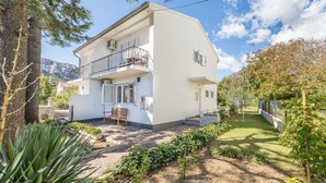 A two-story white building with a balcony, a stone patio, and a garden with plants and a bench. - Apartment Jadranka with garden and barbeque (Baška)