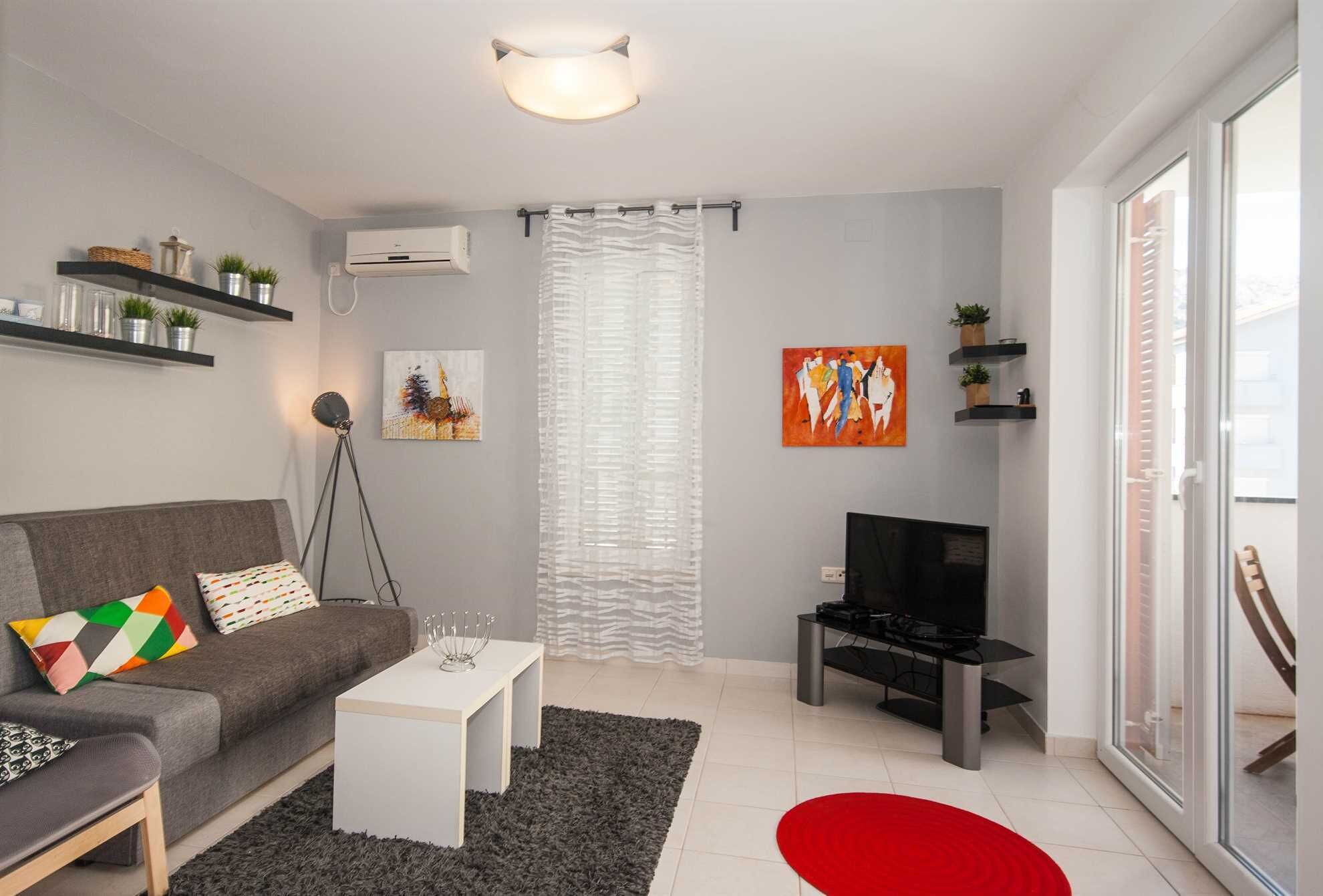 A modern living room with a grey sofa, a white coffee table, a black TV stand, and a red rug. There are shelves with plants and artwork on the walls.