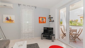 A modern living room with a flat-screen TV, a grey sofa, a coffee table, and a red rug. There is a sliding glass door leading to an outdoor area with a table and chairs. - Vera - Cozy Solo Retreat: 1BR Apartment for Two, Parking Included (Baška)