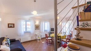 A living room with a sofa, a dining table with chairs, a TV, and a staircase with a decorative item. - Holiday apartment Tinka in centre of Baska with seaview (Baška)