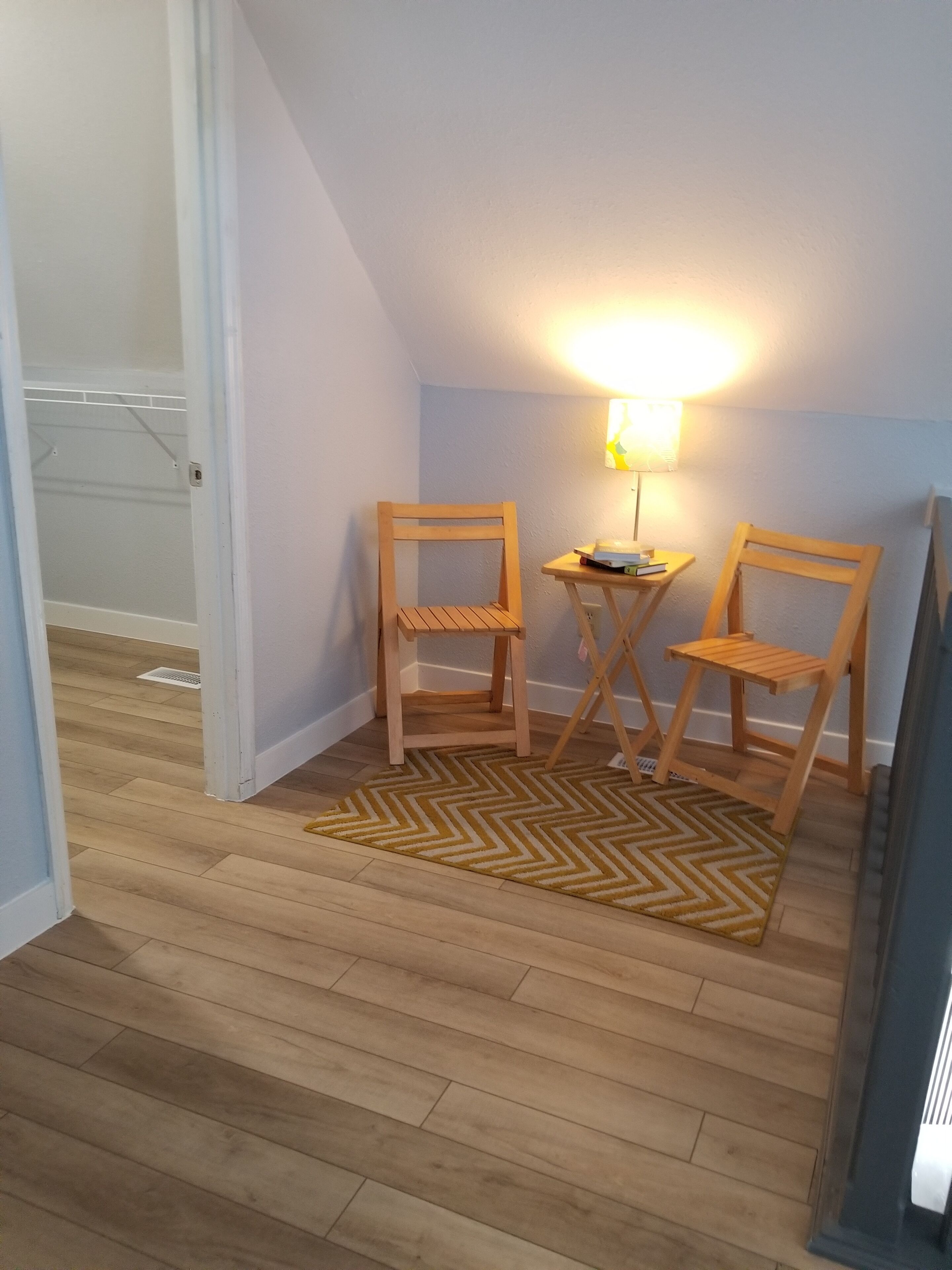 A small room with two wooden chairs, a small table, and a lamp. There is a wooden floor and a door leading to another room.