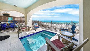 A pool area with a view of the ocean, lounge chairs, and a table.