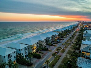 A coastal hotel with multiple-story buildings, a beachfront, and a road lined with palm trees.