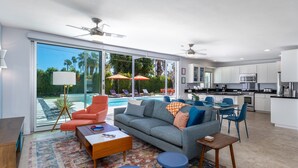 Un salon moderne avec un canapé, une table basse et des fauteuils, une cuisine avec des meubles blancs et un espace piscine visible à travers des portes vitrées.
