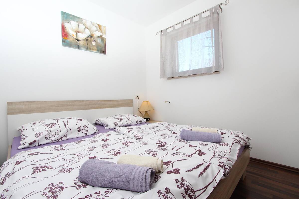 Una cama bien hecha con ropa de cama floral, una lámpara de noche y una ventana con cortina translúcida.