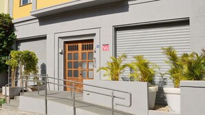 Un edificio con fachada gris, una puerta de madera con paneles de vidrio y una escalera metálica que lleva a la entrada.