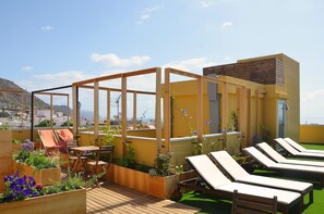 A rooftop terrace with wooden lounge chairs, a wooden table, and planters with flowers.