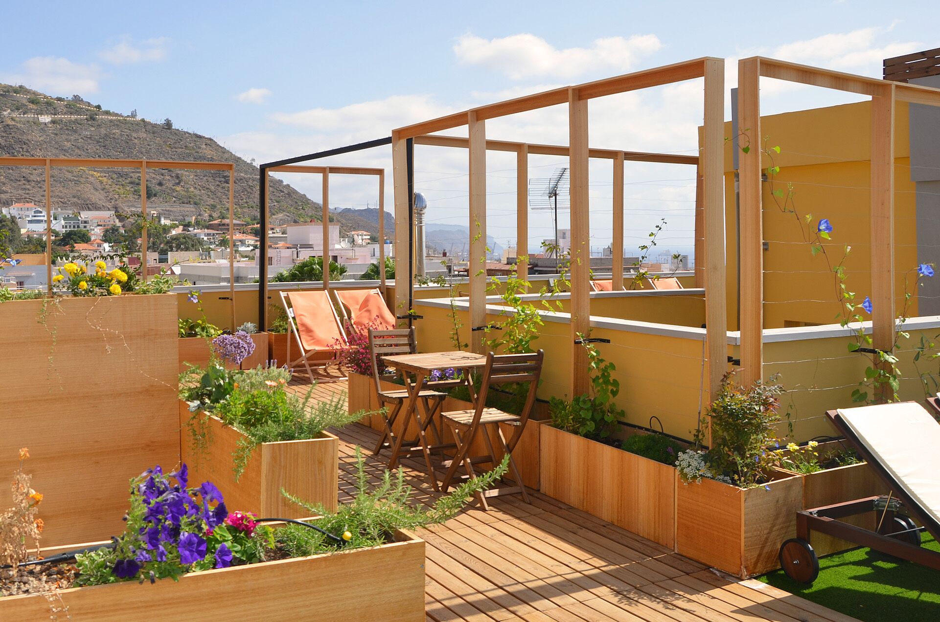 Un jardín en la azotea con terraza de madera, zona de estar y macetas elevadas con flores.