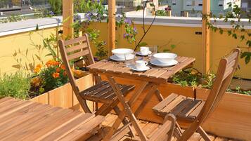 A wooden table set with white plates, cups, and glasses, surrounded by wooden chairs on a balcony with yellow walls and plants.