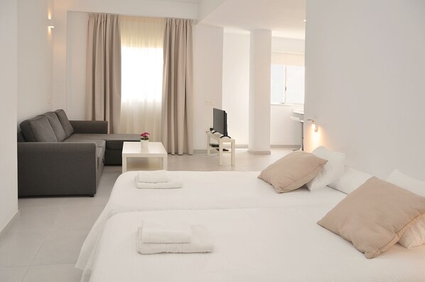 A modern hotel room with a sofa, a bed with two pillows, a small table with a TV, and a window with curtains.