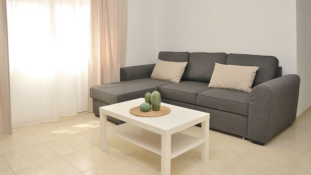 Un salon avec un canapé gris, deux coussins beige, une table basse blanche ornée d’un chemin de table tissé et de deux cactus verts, ainsi qu’une fenêtre avec des rideaux légers.