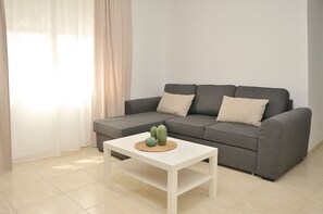 Sala de estar com sofá cinza, duas almofadas bege, mesa de centro branca com um descanso de palha e dois cactos verdes, além de uma janela com cortinas leves.