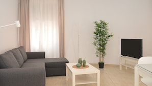 Ein modernes Wohnzimmer mit einem grauen Sofa, einem weißen Couchtisch mit Dekoration, einem Blumentopf, einem Flachbildfernseher auf einem Standfuß und einer Lampe.