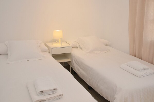 A hotel room with two single beds, a nightstand with a lamp, and a folded towel on each bed.