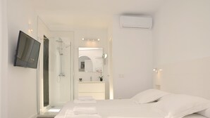 Ein modernes Hotelzimmer mit einem großen Bett, einem an der Wand montierten Fernseher, einer Duschkabine und einem Badezimmer mit Spiegel und Waschbecken.