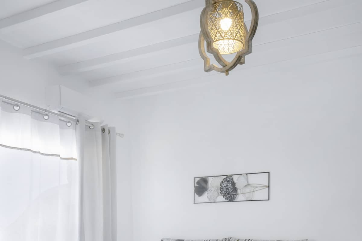 A white bedroom with a bed, a hanging light fixture, a window with curtains, and a wall-mounted artwork.