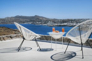 Zwei weiße Stühle und ein kleiner Tisch mit zwei Orangensäften und einer Speiseplatte auf einer Terrasse mit Blick auf eine Küstenlandschaft und Berge im Hintergrund.