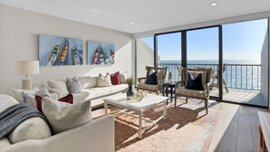 Un salón moderno con un gran ventanal con vistas al mar, un sofá blanco, una mesa de centro y un balcón con mobiliario exterior.