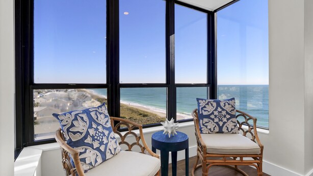 Una habitación con dos sillones de mimbre con cojines blancos y almohadas estampadas en azul, una mesita redonda con un objeto blanco, y un amplio ventanal que ofrece vistas a la playa y al océano.
